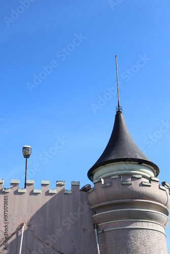 Quadro su tela ​An old, weathered castle wall with battlements and a round turret topped by a conical black spire stands against a clear, deep blue sky during daytime, showing architectural detail