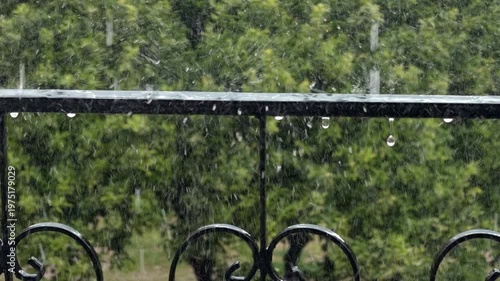 Close Up Of Raindrops Splashing On A Black Metal Balcony Fence During A Summer Storm. Lush Green Trees In The Blurry Background Create A Peaceful And Moody Rainy Day Atmosphere.