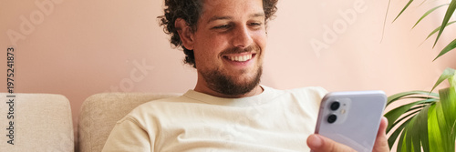 Positive young man with curly hair and beard, sitting on sofa in cozy living room, using mobile phone. Panoramic