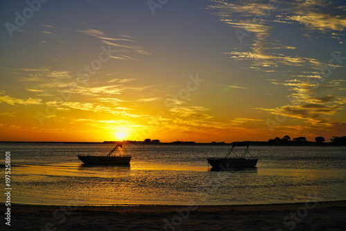 Two small boats silhouetted at golden sunset over calm coastal waters with sandy beach