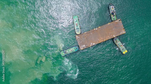 Four tugboats working together to maneuver a massive industrial metal barge in the open sea. A high-angle view showcasing maritime logistics, cooperative teamwork, and heavy cargo transportation.