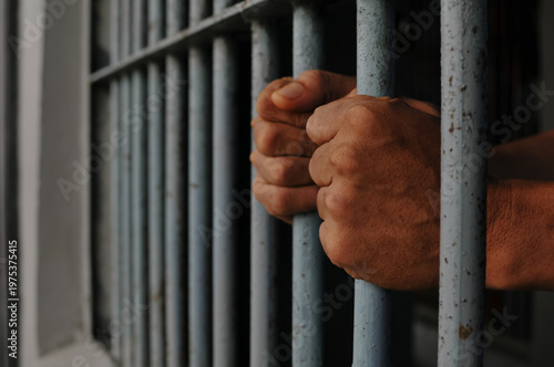 Fotografía Close up of a prisoner hand gripping rusty iron bars behind a jail cell or detention center