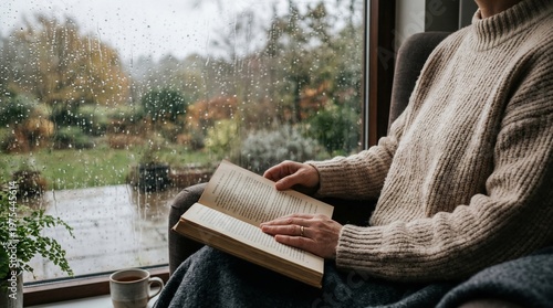Woman reading book on rainy day by window, relaxing with cozy sweater and garden view
