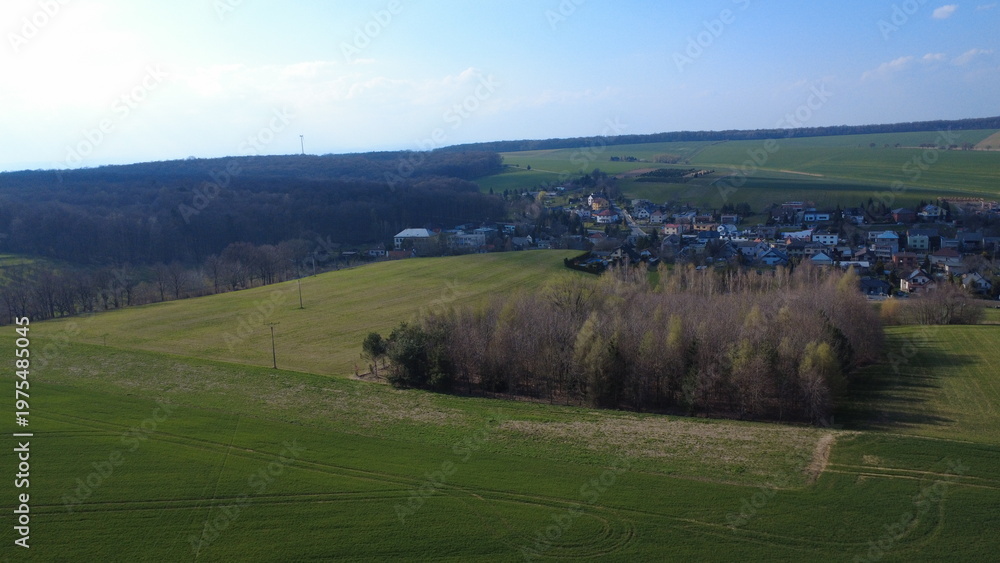 Fototapeta premium Aerial drone view of Hněvošice in the Czech Republic during a sunny spring day. The image shows a peaceful village surrounded by green fields under a clear blue sky, capturing the beauty of the Czech 