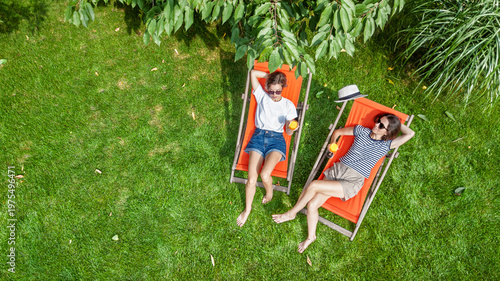Young girls relax in summer garden in sunbed deckchairs on grass, women friends having drinks outdoors in green park lawn on weekend, aerial drone view 