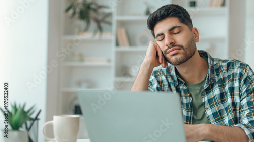 Φωτογραφία A young man sits at a desk with his hand on his face, appearing tired and distracted