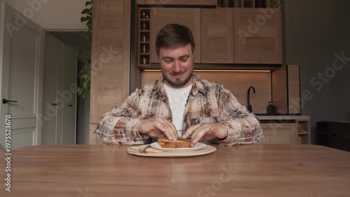 A man is sitting at home at a table in his home clothes and eating a sandwich.