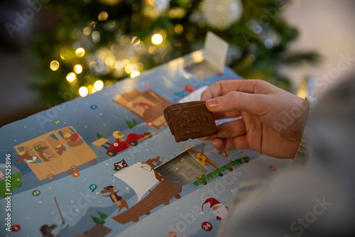 Gros plan d'un enfant ouvrant la fenêtre d'un calendrier de l'Avent avec un chocolat, sapin de noël avec guirlande lumineuse en fond.