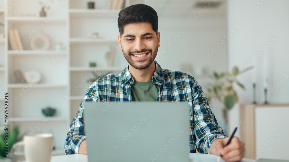 Fototapeta premium A man sits at a table with a laptop, smiling while he works. The room is bright, featuring plants and books on shelves. He holds a pen in his hand, focused on the screen.