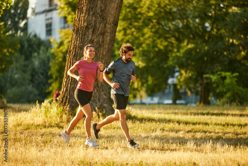 In a vibrant park, a couple jogs happily side by side on grassy terrain during late afternoon. They embrace a healthy lifestyle and enjoy the beautiful weather.