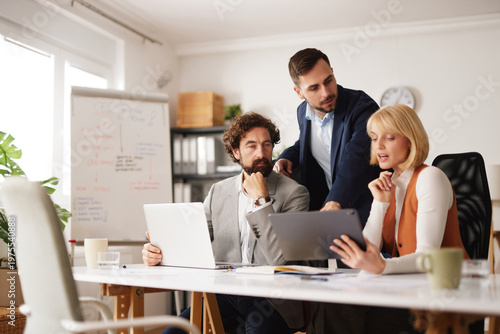 A business team works together in an office. They gather around a table focusing on a tablet while discussing ideas and plans. Laptops and notes are present as they plan for upcoming projects.