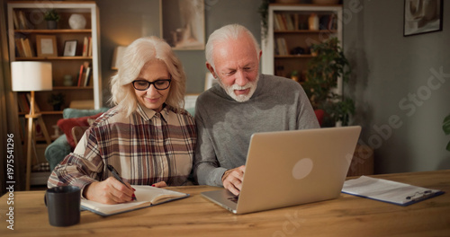 A couple is seated at a wooden table. They are focused on a laptop while taking notes in a notebook. Bookshelves and a plant are visible in the background.