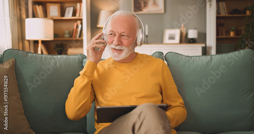 An elderly man enjoys his time on the couch. He wears headphones and talks on the phone while holding a tablet. The living room has a cozy setup with soft furniture and warm lighting.
