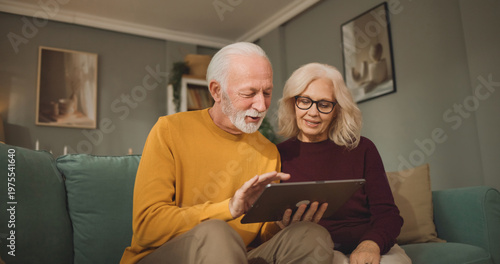 An elderly couple sits on a couch in their living room, looking at a tablet to explore life insurance options and discuss financial planning together during the evening hours.
