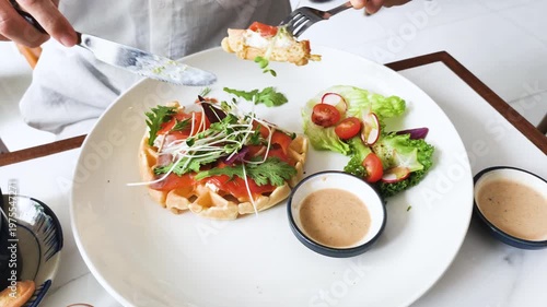 Hands dip elegant salmon for waffle brunch into creamy sauce. Person uses fork to soak piece in dressing. High angle shows elegant salmon and waffle brunch with dip. Fresh taste is captured.
