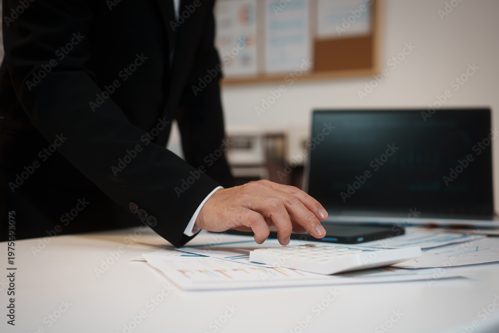 Fototapeta premium Businessman in suit using calculator on financial reports at office desk with laptop in background, analyzing budget and accounting data for corporate finance.