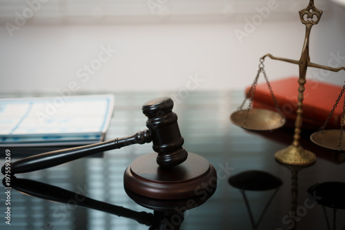 Fotografie Close-up of wooden judge gavel on reflective desk with brass scales of justice and law books in background, shallow depth of field, legal concept
