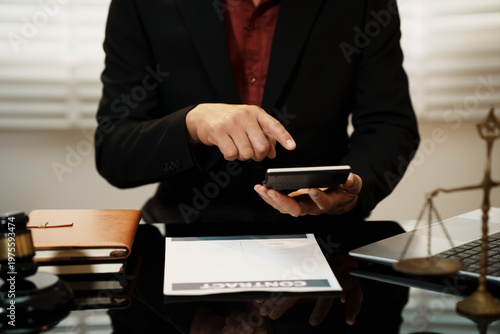Business professional in suit using tablet at office desk with contract papers, gavel, laptop and justice scales, suggesting legal consultation and digital law services.