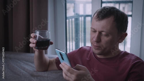 Portrait of a young, attractive man browsing messages on his phone and drinking from a glass while sitting on a sofa.