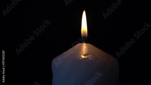 Single candle flame glows softly against black background. Candle wax melts gently around the wick's base. Light flickers, casting warm halo around the flame