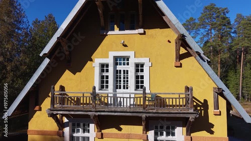 Yellow house with wooden balcony and white windows surrounded by trees, showcasing a clear blue sky and natural landscape in the background