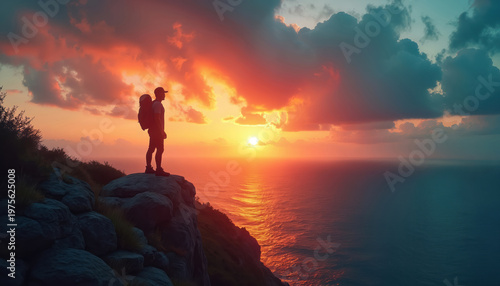 Man stands on cliff edge watching sunset over ocean. Silhouette backpacker on mountain peak admires twilight sky. Coastal view, sea horizon, warm sky colors.