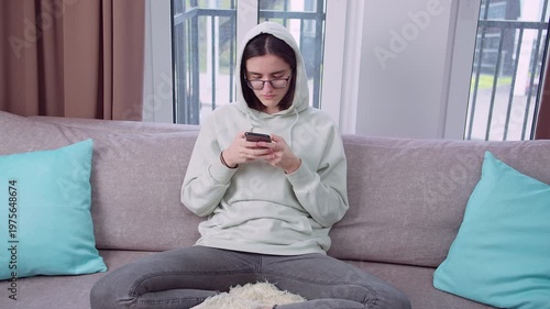 Young brunette woman in a hoodie and glasses sitting alone on a sofa, reading messages on her smartphone. Front view at eye level, daylight from window, serious and sad emotional mood.