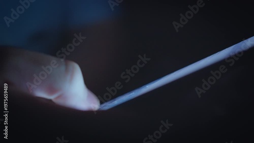 Close-up detail of a male thumb scrolling on a mobile phone in the dark. The light from the screen illuminates the hand, capturing a calm, modern moment of online connection and everyday technology.