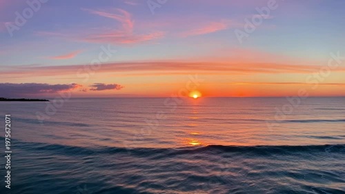 Vibrant sunset over the calm ocean, golden sun sitting on the horizon with soft pink and orange clouds in the sky, peaceful seaside landscape.