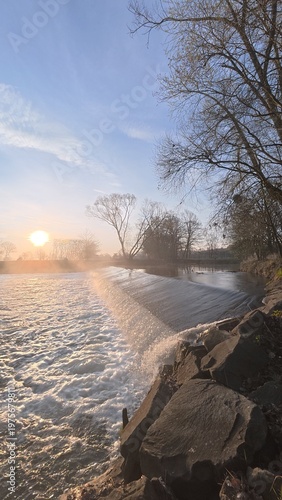 Morning weir on the Opava River at sunrise, with soft mist rising above the water and gentle light illuminating the peaceful river landscape. The scene captures calm morning nature atmosphere