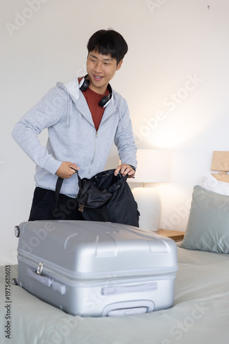 Asian man packing black backpack beside bed, glancing at silver suitcase, wearing hoodie
