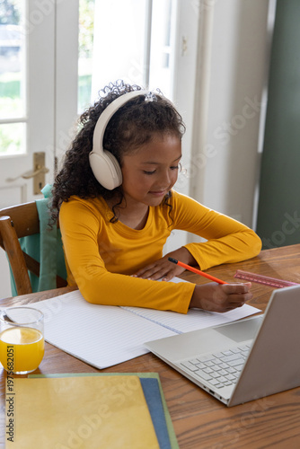 African American child studying at wooden table near glass doors with laptop headset notebook