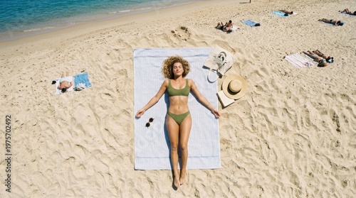 Aerial view of a smiling young woman in a green bikini relaxing and sunbathing on a towel on a beautiful sandy beach by the turquoise ocean during summer vacation.