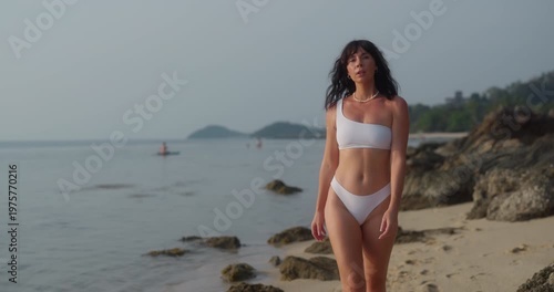 Portrait of thoughtful sportive woman in swimwear walks along sea beach at sunset. Slim female looks at tranquil water of ocean as rests at exotic resort
