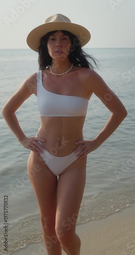 Portrait of pretty woman in bikini and straw hat puts hands on hips on sea beach vertical shot. Attractive slim lady enjoys calm day on azure ocean coast