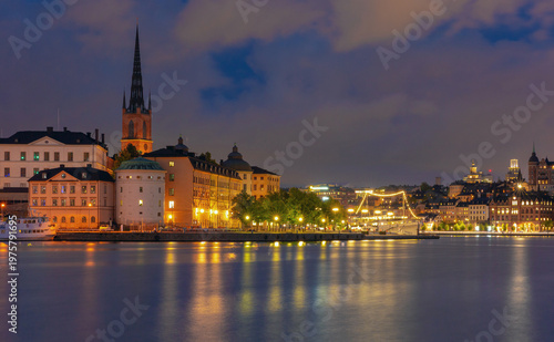 Blue hour view of Stockholm waterfront in Sweden with illuminated historic buildings, church spire and reflections on calm water.