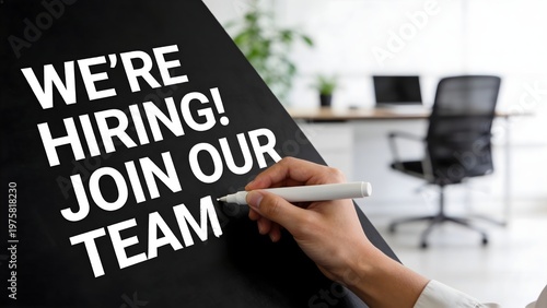 Person uses a white marker to write we are hiring join our team on a large black board surface display