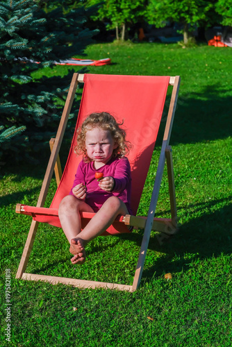 Moscow, Russia, 23.03.2026. Young girl crying with strawberry while sitting on red deck chair on green grass in summer garden. Childhood emotions at dacha.
