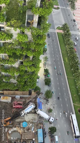 Aerial view of a construction site with green walls, trucks, and a busy road. Trees and plants are visible on the building facade.