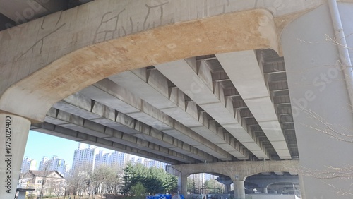 Underneath a massive concrete bridge structure with repetitive beams and city view in background