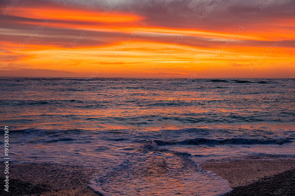 Fototapeta premium Dramatic Golden sunset over tranquil beach, waves lashing the pebble coastline