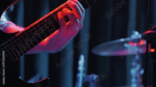 Close-up of a guitarist's hand on a fretboard playing fast. Dramatic purple stage lighting creates an energetic mood. Focus on the musician, with a blurred drum kit in the background