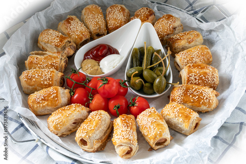 Hot Dog style sausages baked in puff pastry, sprinkled with sesame seeds, with capers, pickles and cherry tomatoes.