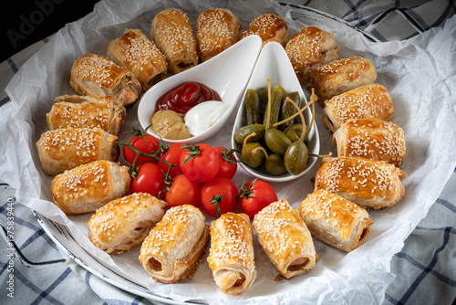 Hot Dog style sausages baked in puff pastry, sprinkled with sesame seeds, with capers, pickles and cherry tomatoes.