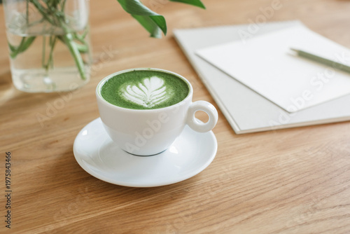 Creative desk setup featuring a hot matcha drink, blank paper mockup, spiral notebook, and flowers on a wooden table.