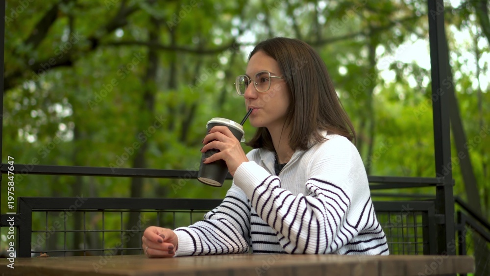Fototapeta premium Young woman in a white sweater sipping coffee on a cafe veranda by the forest