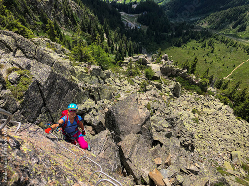 Young adult woman equipped with helmet and harness enjoying a thrilling adventure, climbing a rocky via ferrata path with metal rungs and safety cables high above a deep valley, Chamonix, France.