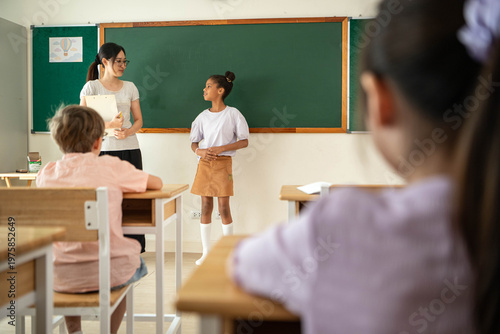 Schoolgirl presenting in front of class with teacher observing