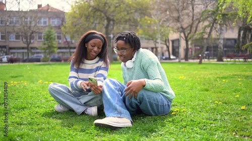 Smiling friends sitting on the grass and using a smartphone