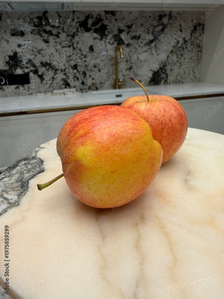 Obraz premium Two apples with red and yellow hues rest on a round, creamy marble surface. The background features a modern kitchen with a gold faucet and a black-white backsplash